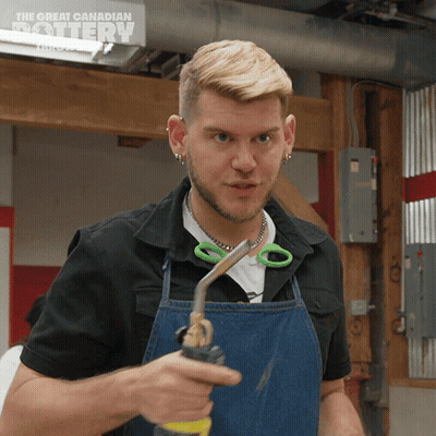 A Man Wearing An Apron Is Holding A Pair Of Scissors In Front Of A Sign That Says The Great Canadian Pottery GIF