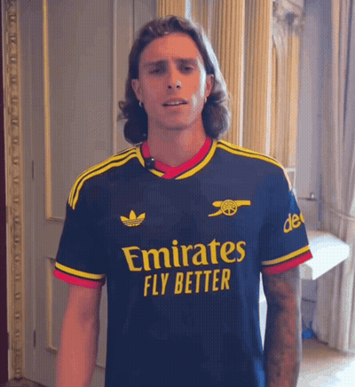 A Man Wearing An Emirates Fly Better Shirt Stands In A Room GIF