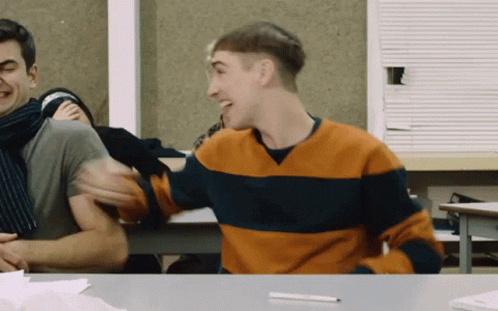 A Man Wearing An Orange And Blue Striped Sweater Laughs While Sitting At A Desk GIF