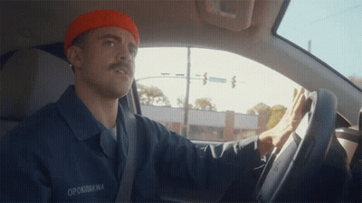 A Man Wearing An Orange Beanie And A Blue Shirt With The Name Opokukwa On It Is Driving A Car GIF