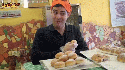 A Man Wearing An Orange Hat Is Sitting At A Table With A Bunch Of Donuts GIF