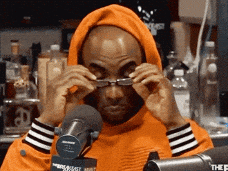 A Man Wearing An Orange Hoodie Is Adjusting His Glasses In Front Of A Microphone That Says Breakfast Adult GIF