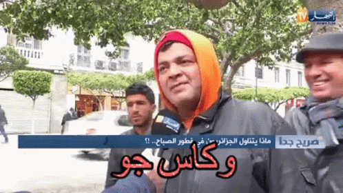 A Man Wearing An Orange Hoodie Is Talking Into A Microphone While Another Man Stands Behind Him GIF