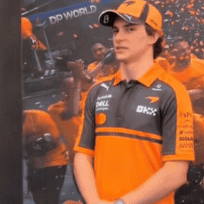 A Man Wearing An Orange Shirt And A Hat Is Standing In Front Of A Picture Of A Crowd . GIF