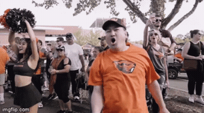 A Man Wearing An Orange Shirt With A Buffalo Bills Logo Stands In Front Of A Cheerleader GIF