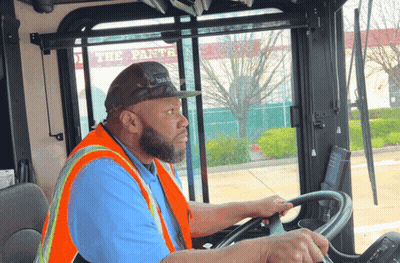 A Man Wearing An Orange Vest And A Hat Is Driving A Bus GIF