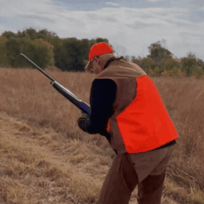 A Man Wearing An Orange Vest Is Holding A Shotgun In A Field GIF