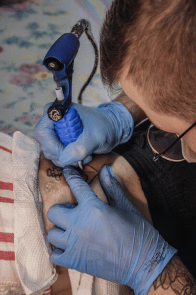 A Man Wearing Blue Gloves Is Getting A Tattoo On Someone 'S Leg GIF