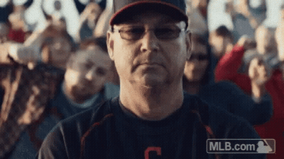 A Man Wearing Glasses And A Baseball Cap Is Sitting In Front Of A Crowd Of People . GIF