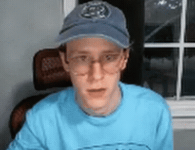 A Man Wearing Glasses And A Baseball Cap Is Sitting In Front Of A Window . GIF