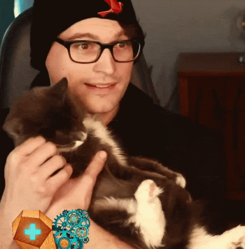 A Man Wearing Glasses And A Black Hat Holds A Cat In His Arms GIF