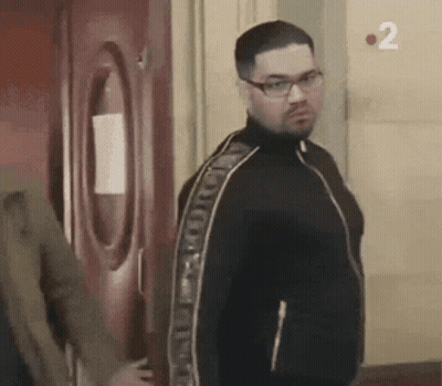 A Man Wearing Glasses And A Black Jacket Is Standing In Front Of A Door With The Number 2 On It . GIF