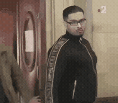 A Man Wearing Glasses And A Black Jacket Is Standing In Front Of A Red Door . GIF
