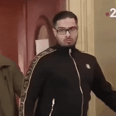 A Man Wearing Glasses And A Black Jacket Is Standing Next To A Door . GIF