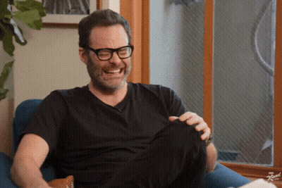 A Man Wearing Glasses And A Black Shirt Is Sitting In A Chair Laughing GIF