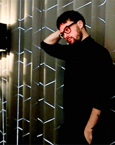 A Man Wearing Glasses And A Black Shirt Is Standing In Front Of A Metal Wall GIF
