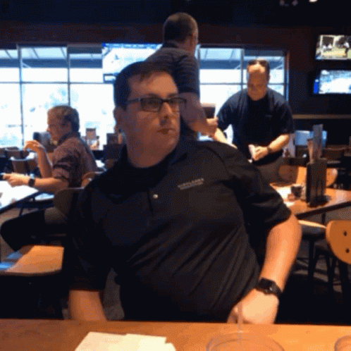 A Man Wearing Glasses And A Black Shirt That Says ' Oklahoma ' On It Sits At A Table In A Restaurant GIF