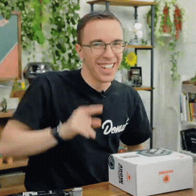 A Man Wearing Glasses And A Black Shirt With The Word Donut On It Is Sitting At A Table With A Box On It . GIF