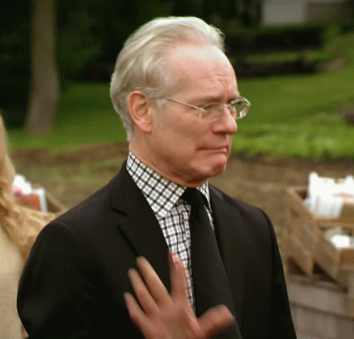 A Man Wearing Glasses And A Black Suit Stands In Front Of A Green Field GIF