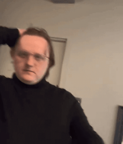 A Man Wearing Glasses And A Black Turtleneck Is Holding His Hair . GIF