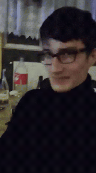 A Man Wearing Glasses And A Black Turtleneck Is Smiling In Front Of A Coke Bottle GIF