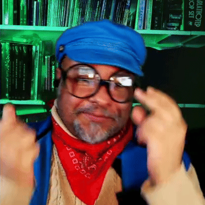 A Man Wearing Glasses And A Blue Hat Is Standing In Front Of A Bookshelf That Has A Book Titled Blastworld On It GIF