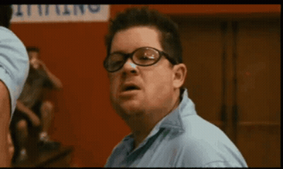 A Man Wearing Glasses And A Blue Shirt Holds Up His Fist In Front Of A Sign That Says Wrestling GIF