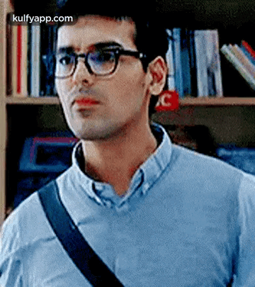A Man Wearing Glasses And A Blue Shirt Is Standing In Front Of A Bookshelf In A Library . GIF