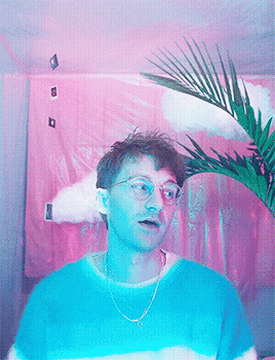 A Man Wearing Glasses And A Blue Shirt Stands In Front Of A Pink Curtain GIF