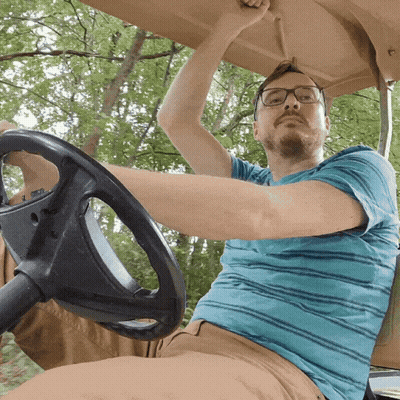 A Man Wearing Glasses And A Blue Striped Shirt Is Driving A Golf Cart GIF