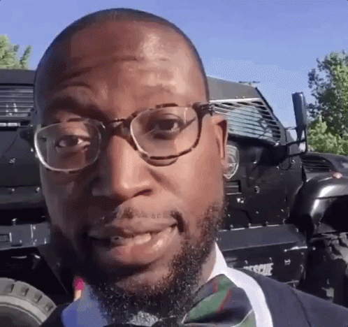A Man Wearing Glasses And A Bow Tie Is Smiling In Front Of A Vehicle . GIF