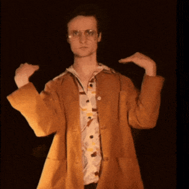 A Man Wearing Glasses And A Brown Coat Is Holding His Hands Up In The Air GIF