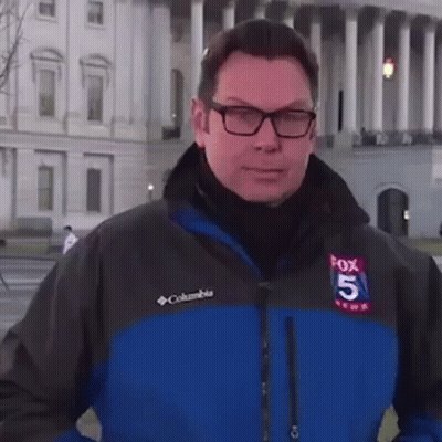 A Man Wearing Glasses And A Columbia Jacket Is Standing In Front Of A Large Building . GIF