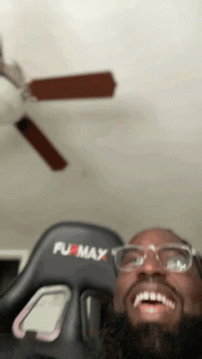 A Man Wearing Glasses And A Furmax Gaming Chair Is Smiling In Front Of A Ceiling Fan . GIF