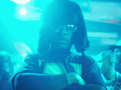 A Man Wearing Glasses And A Furry Hat Is Standing In Front Of A Crowd Of People GIF