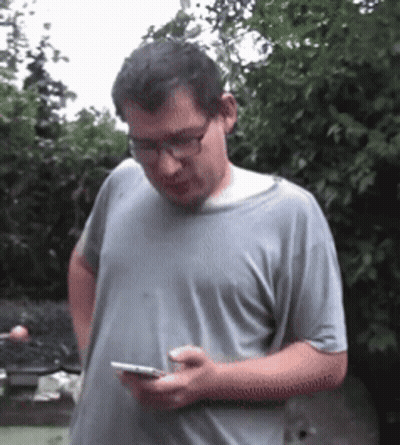 A Man Wearing Glasses And A Gray Shirt Is Looking At His Phone GIF