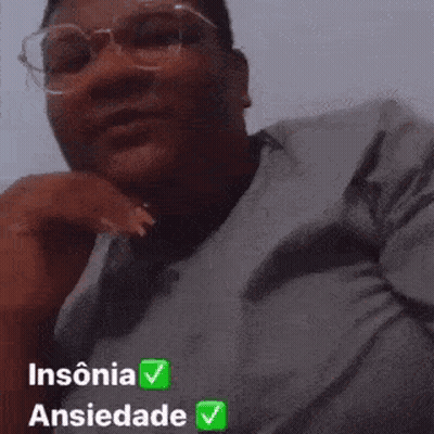 A Man Wearing Glasses And A Gray Shirt Is Sitting Down With His Hand On His Chin And Looking At The Camera . GIF