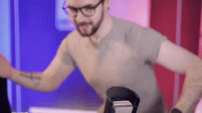 A Man Wearing Glasses And A Gray Shirt Is Standing In Front Of A Microphone . GIF