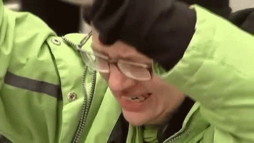 A Man Wearing Glasses And A Green Jacket Is Smiling While Holding His Head . GIF