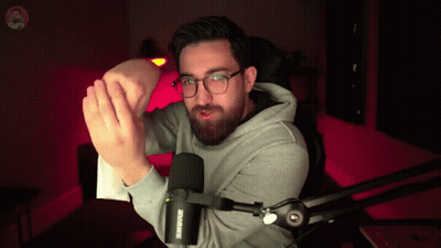 A Man Wearing Glasses And A Grey Hoodie Is Standing In Front Of A Shure Microphone GIF