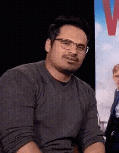 A Man Wearing Glasses And A Grey Shirt Is Sitting In Front Of A Sign . GIF
