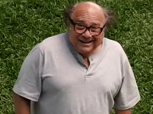 A Man Wearing Glasses And A Grey Shirt Is Standing In The Grass . GIF