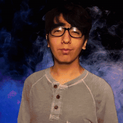 A Man Wearing Glasses And A Grey Shirt Stands In Front Of Smoke GIF