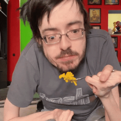 A Man Wearing Glasses And A Grey Shirt With The Letter S On It Is Eating Food GIF