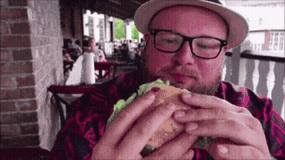 A Man Wearing Glasses And A Hat Is Eating A Burger GIF