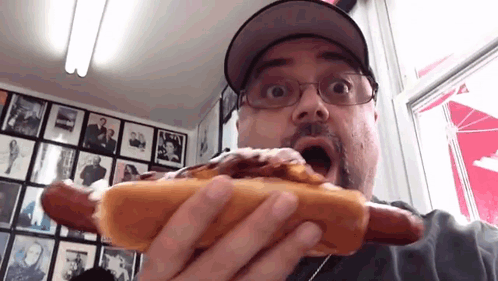 A Man Wearing Glasses And A Hat Is Eating A Hot Dog With A Surprised Look On His Face GIF