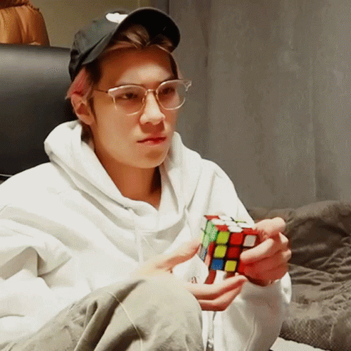 A Man Wearing Glasses And A Hat Is Holding A Rubik 'S Cube In His Hands GIF