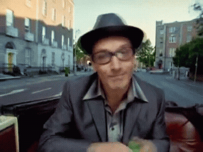 A Man Wearing Glasses And A Hat Is Sitting On A Red Couch GIF