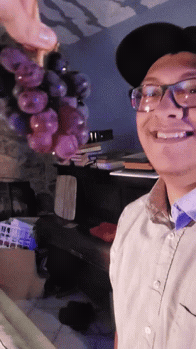 A Man Wearing Glasses And A Hat Is Smiling While Holding A Bunch Of Grapes GIF