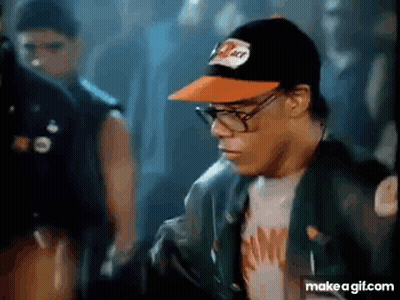 A Man Wearing Glasses And A Hat Is Standing In A Crowd Of People . GIF